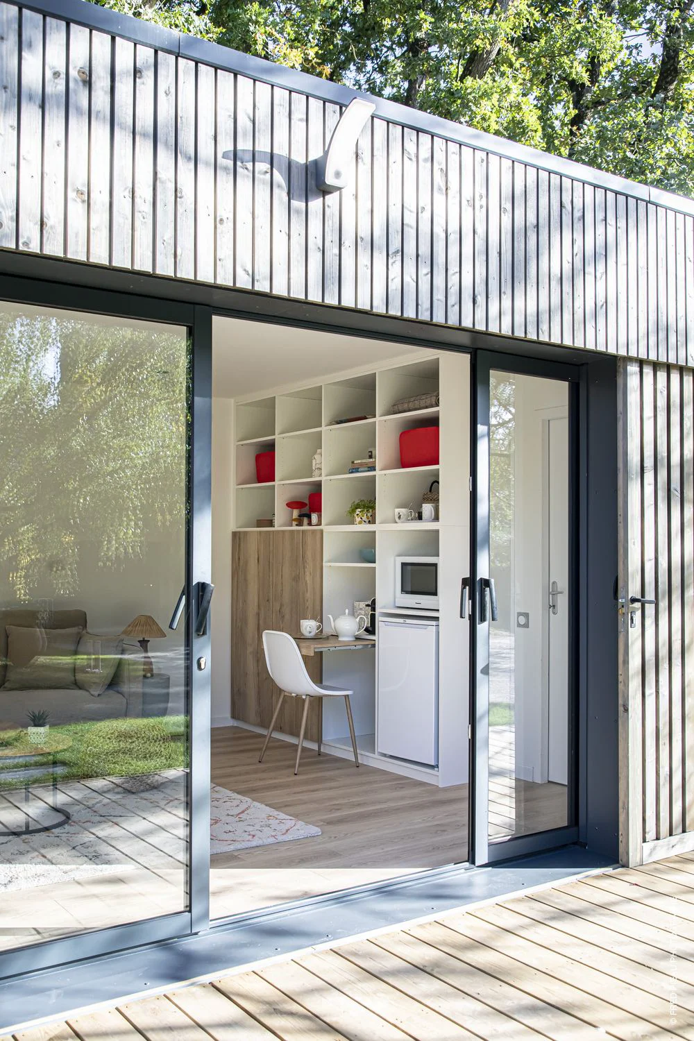 Intérieur d’un espace compact en bois avec kitchenette, étagères intégrées et coin salon visible depuis des portes vitrées ouvertes.