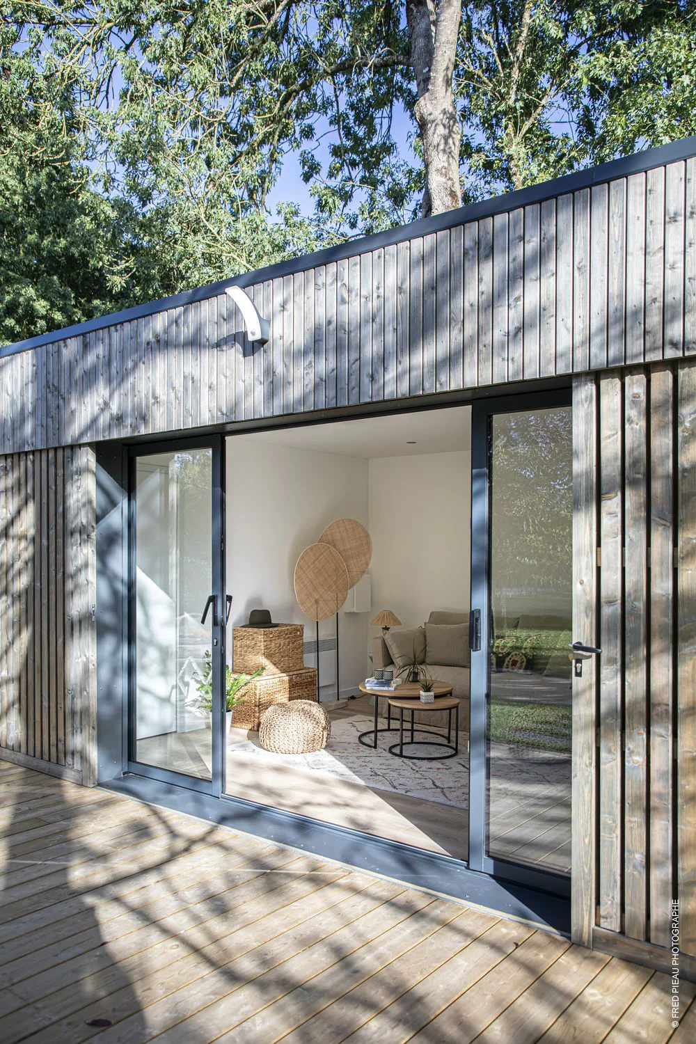 Bâtiment moderne habillé de bois avec portes vitrées ouvertes, terrasse et intérieur lumineux avec espace détente.